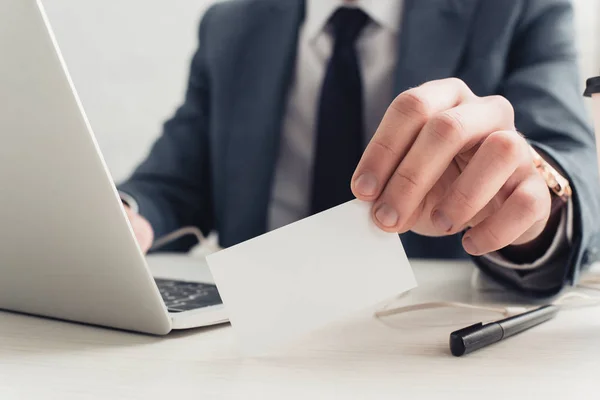 Cropped view of businessman holding blank business card while sitting at workplace near laptop — Stock Photo