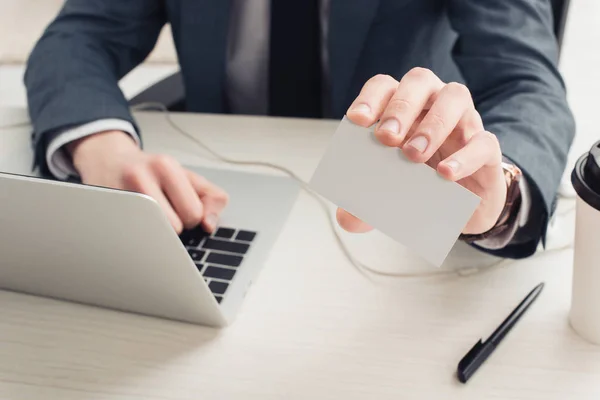 Partial view of businessman holding empty business card while sitting near laptop — Stock Photo