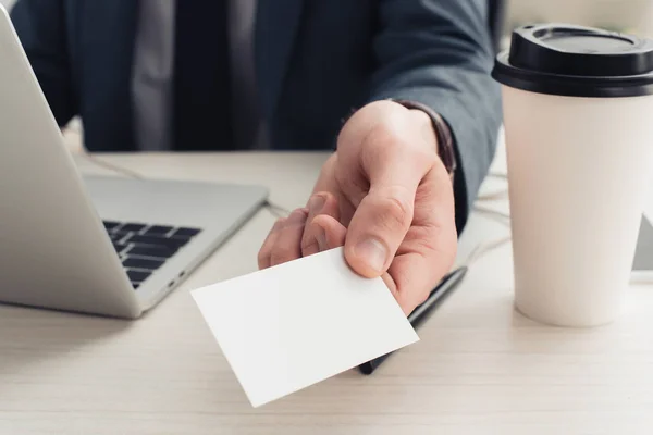 Cropped view of businessman holding empty business card while sitting near laptop and coffee to go — Stock Photo