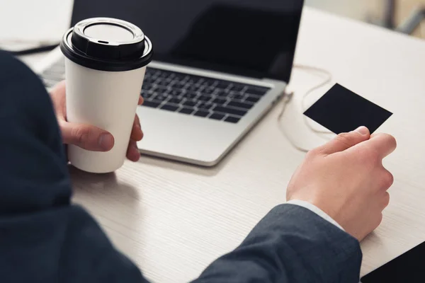 Vista recortada de hombre de negocios sosteniendo café para ir y vacío tarjeta de visita negro mientras está sentado en el lugar de trabajo cerca de la computadora portátil - foto de stock