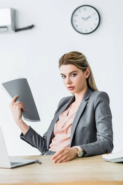Mujer de negocios sentada en la oficina con la carpeta en la mano, mirando a la cámara y sufriendo de calor - foto de stock