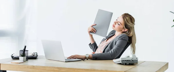 Panoramic shot of businesswoman sitting in office, holding folder in hand and suffering from heat — Stock Photo