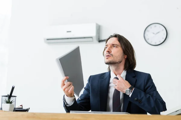 Hombre de negocios sentado en la oficina con la carpeta en la mano, mirando hacia otro lado y sufriendo de calor - foto de stock