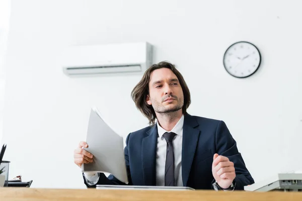 Hombre de negocios sentado en la oficina, sosteniendo la carpeta en la mano, mirando hacia otro lado y sufriendo de calor - foto de stock
