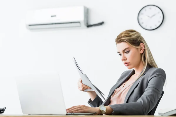 Mujer de negocios sentada en la oficina con aire acondicionado, la celebración de la carpeta en la mano y el uso de ordenador portátil - foto de stock