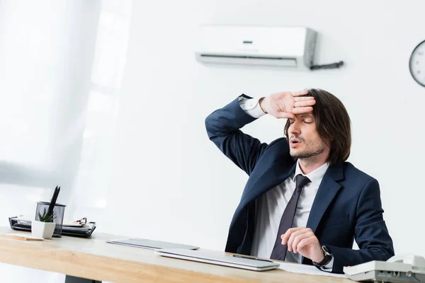 Homme d'affaires en sueur assis au bureau, tenant la main près du visage et souffrant de chaleur — Photo de stock