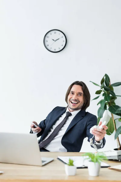 Happy businessman holding headset and smartphone in hands, sitting behind table and looking at camera — Stock Photo