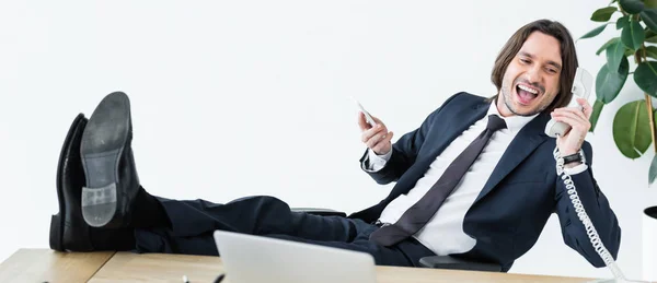 Panoramic shot of cheerful businessman talking at telephone, holding smartphone and headset in hands — Stock Photo