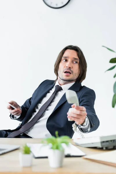 Handsome businessman holding headset and smartphone in hands, sitting behind table and looking away — Stock Photo