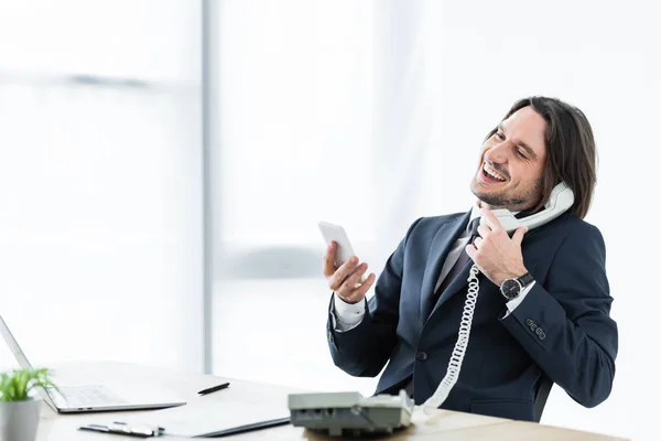 Cheerful businessman talking at telephone, holding smartphone and headset in hands — Stock Photo