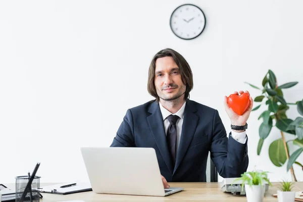 Businessman holding red heart in hand, sitting in office and looking at camera, medical insurance concept — Stock Photo