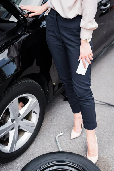 Cropped view of businesswoman with smartphone in hand standing near broken auto, car insurance concept — Stock Photo
