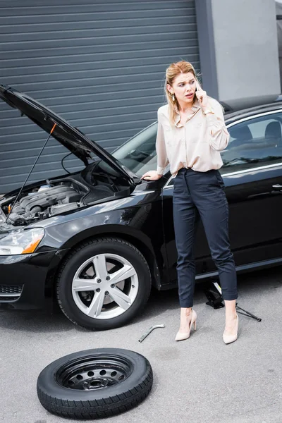 Mujer de negocios mirando hacia otro lado, hablando en el teléfono inteligente y de pie cerca de auto roto, concepto de seguro de coche - foto de stock