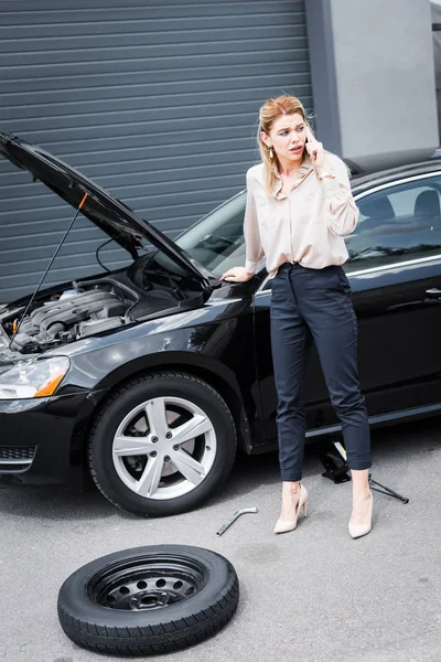 Business woman looking away, talking on smartphone and standing near broken auto, car insurance concept — стоковое фото