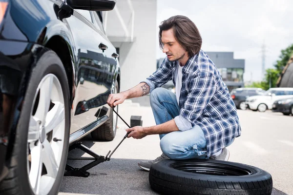 Hombre en el desgaste casual usando la herramienta del gato y cambiando la rueda rota en auto, concepto de seguro de coche - foto de stock