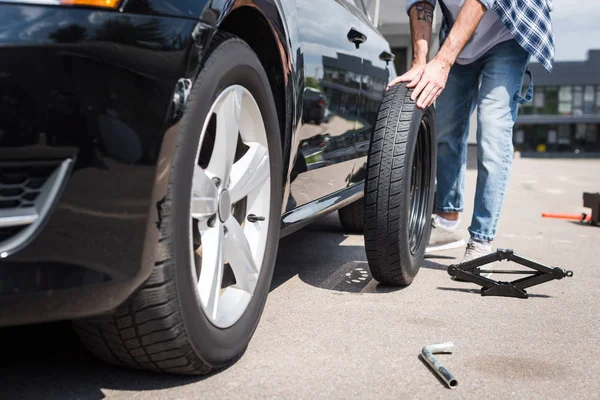 Cropped view of man rolling new tire and fixing broken auto, car insurance concept — Stock Photo