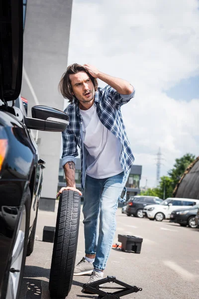 Hombre guapo en ropa casual rodando nueva rueda y la fijación de automóviles rotos, concepto de seguro de coche - foto de stock