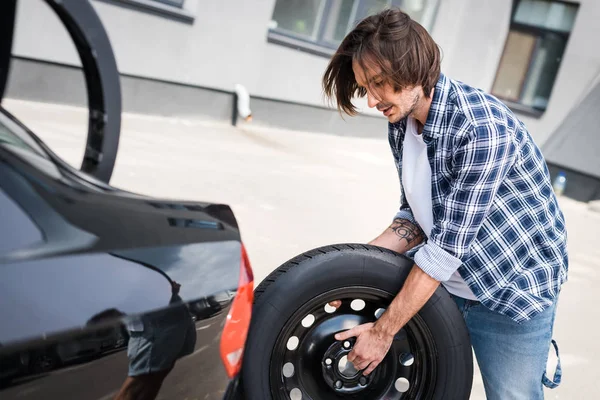 Hombre sosteniendo nueva rueda en las manos cerca de auto, concepto de seguro de coche - foto de stock