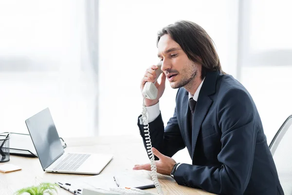 Homme d'affaires parlant au téléphone, assis au bureau et regardant ailleurs — Photo de stock