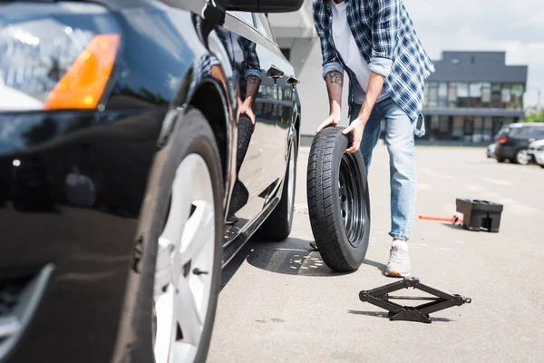 Cropped view of man in casual clothes rolling new wheel and fixing broken auto, car insurance concept — Stock Photo