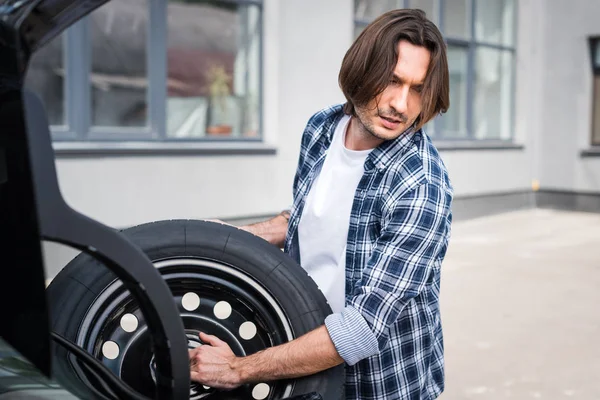 Hombre guapo en ropa casual sosteniendo rueda en las manos cerca de auto, concepto de seguro de coche - foto de stock