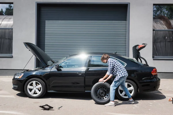 Man rolling new wheel and fixing broken auto near garage, car insurance concept — Stock Photo