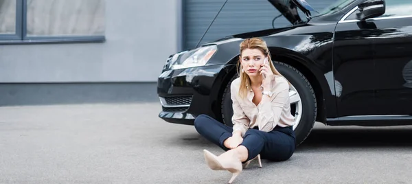 Panoramaaufnahme einer verärgerten Geschäftsfrau, die auf dem Smartphone spricht und in der Nähe eines kaputten Autos sitzt, Kfz-Versicherungskonzept — Stockfoto