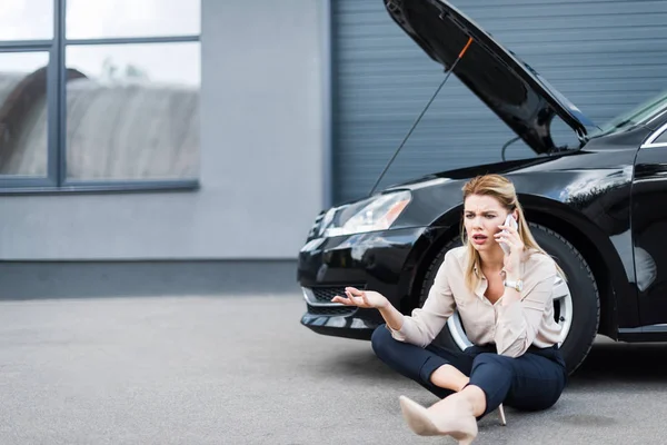 Confusa mujer de negocios hablando en el teléfono inteligente y sentado cerca de auto roto, concepto de seguro de coche - foto de stock