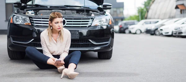 Panoramic shot of upset businesswoman sitting near broken auto with smartphone in hands, car insurance concept — Stock Photo