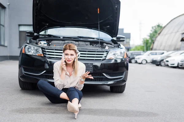 Расстроенная деловая женщина смотрит в сторону, разговаривая на смартфоне и сидя рядом со сломанным авто, концепция страхования автомобиля — стоковое фото