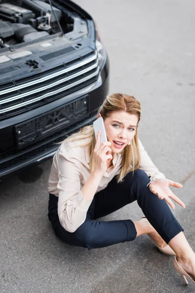 Confused businesswoman looking at camera, talking on smartphone and sitting near broken auto, car insurance concept — Stock Photo