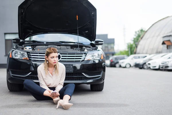 Bouleversé femme d'affaires assis près de cassé auto avec smartphone dans les mains, concept d'assurance automobile — Photo de stock