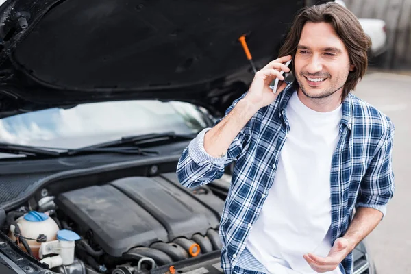 Cheerful man in casual cothes talking on smartphone near broken auto with open trunk, car insurance concept — Stock Photo