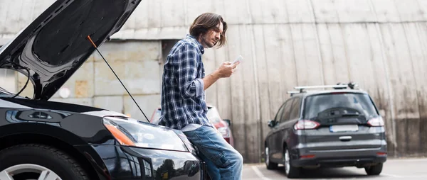 Панорамный снимок человека со смартфоном, стоящим рядом со сломанным автомобилем с открытым багажником, концепция страхования автомобиля — стоковое фото
