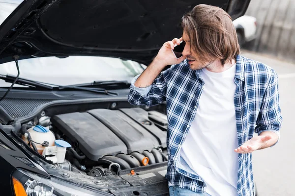 Upset man in casual wear talking on smartphone near broken auto with open trunk, car insurance concept — Stock Photo