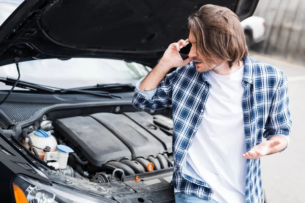 Hombre molesto en ropa casual hablando en el teléfono inteligente cerca de coche roto con maletero abierto, concepto de seguro de coche - foto de stock