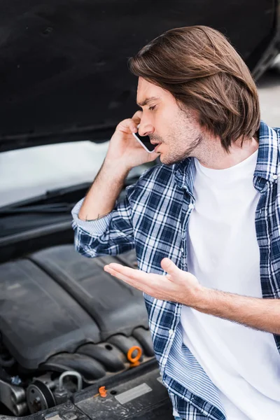 Trauriger und verwirrter Mann spricht auf Smartphone und steht neben kaputtem Auto mit offenem Kofferraum, Kfz-Versicherungskonzept — Stockfoto