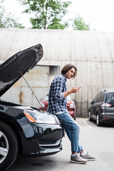 Handsome man with smartphone standing near broken auto with open trunk, car insurance concept — Stock Photo