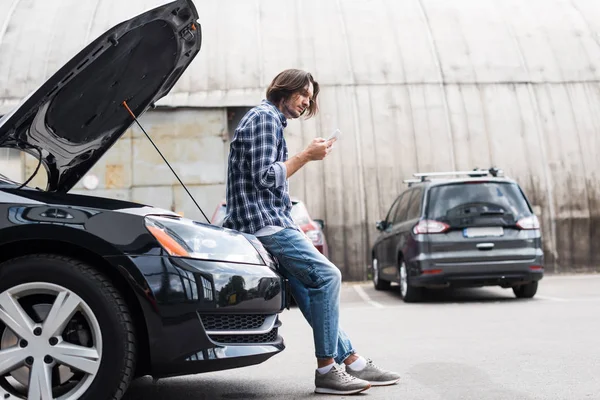 Человек в повседневной одежде со смартфоном в руке, стоящий рядом со сломанным авто с открытым багажником, концепция страхования автомобиля — стоковое фото