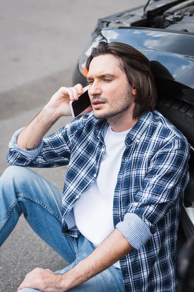 Tired man sitting near broken auto and talking on smartphone, car insurance concept — Stock Photo