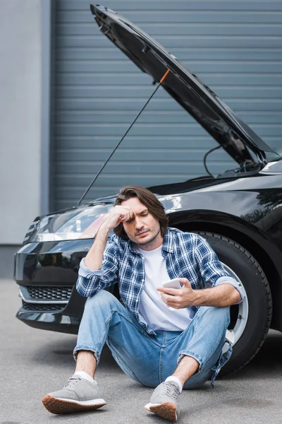 Thoughtful man in casual clothes holding smartphone in hand and sitting near broken auto with open trunk, car insurance concept — Stock Photo