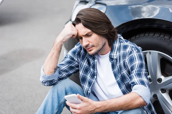 Uomo sconvolto in abiti casual tenendo smartphone in mano e seduto vicino rotto auto con tronco aperto, concetto di assicurazione auto — Foto stock