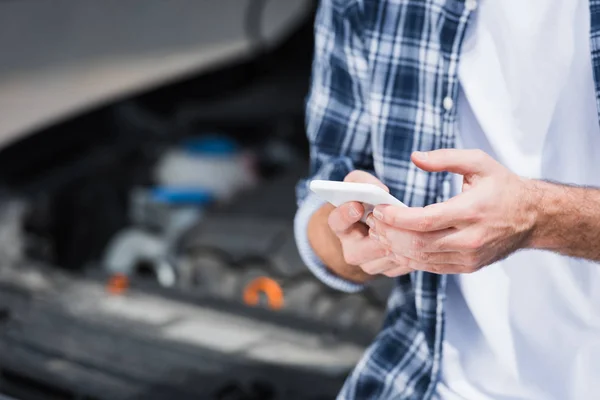 Selective focus of man holding smartphone in hands while standing near broken auto with open trunk, car insurance concept — Stock Photo