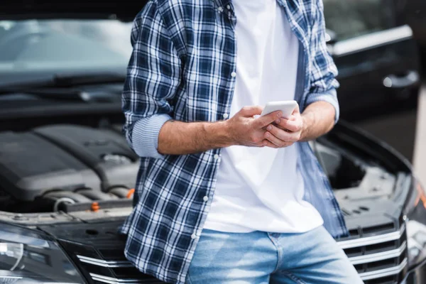 Cropped view of man in casual clothes holding smartphone in hands and standing near broken auto with open trunk, car insurance concept — Stock Photo