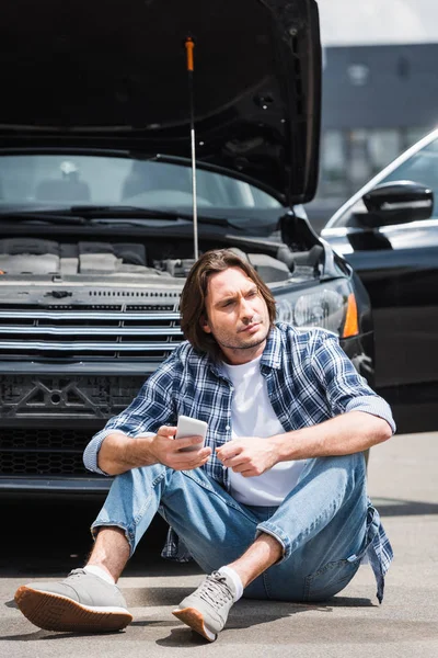 Bell'uomo che tiene lo smartphone in mano, distogliendo lo sguardo e seduto vicino a rotto auto con tronco aperto, concetto di assicurazione auto — Foto stock