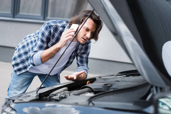 Homem confuso falando no smartphone e de pé perto auto quebrado com tronco aberto, conceito de seguro de carro — Fotografia de Stock