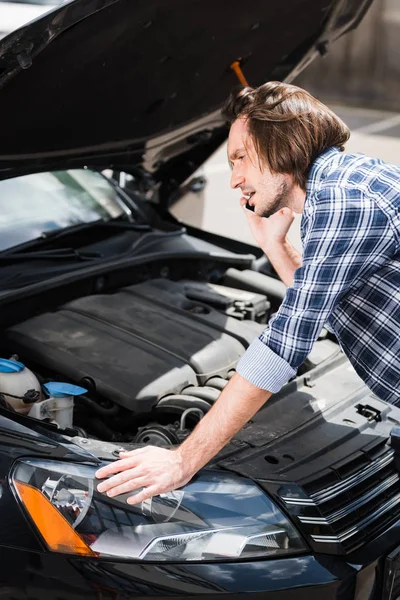 Frustrierter Mann spricht auf Smartphone, während er in der Nähe eines kaputten Autos mit offenem Kofferraum steht, Kfz-Versicherungskonzept — Stockfoto