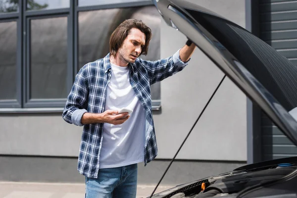 Handsome man holding smartphone in hand and standing near broken auto with open trunk, car insurance concept — Stock Photo