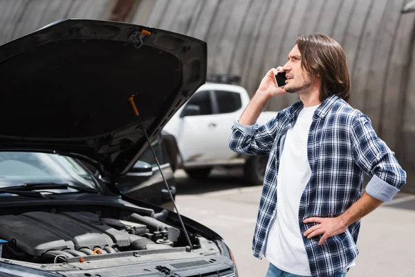 Hombre hablando en el teléfono inteligente, de pie cerca de auto roto con baúl abierto y mirando hacia otro lado, el concepto de seguro de coche - foto de stock