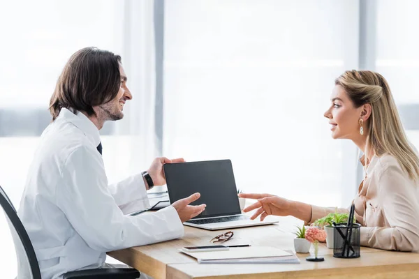 Happy patient looking at doctor and pointing with finger on laptop in hospital — Stock Photo